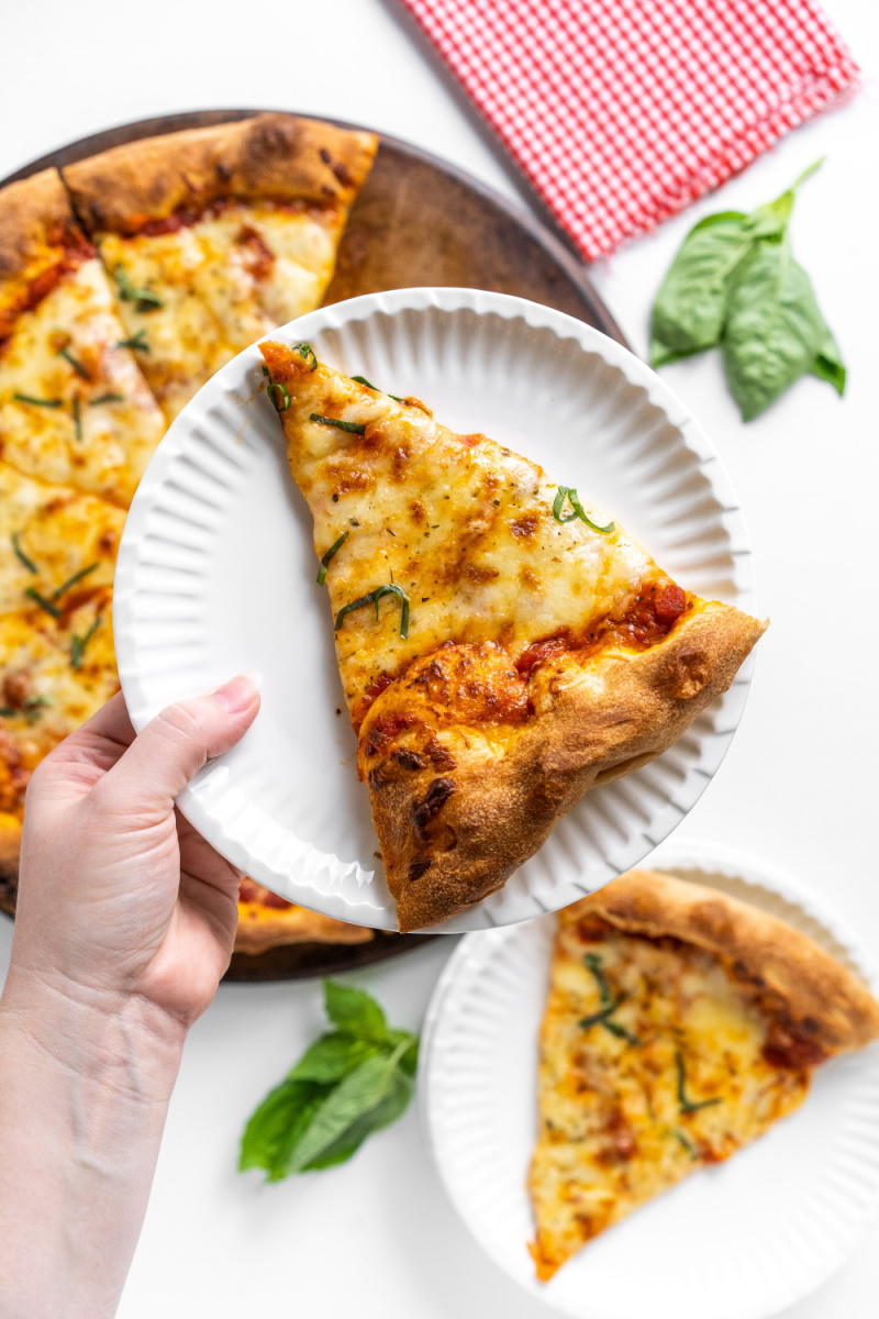 holding slice of cheese pizza on plate over whole pizza